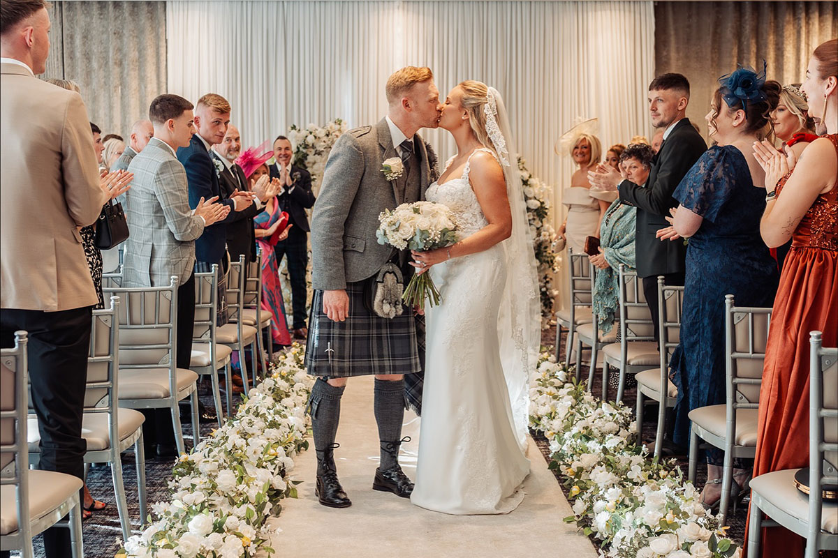 Bride and groom kissing at the end of their wedding ceremony at The Carlton Hotel, with guests applauding and floral arrangements lining the aisle.