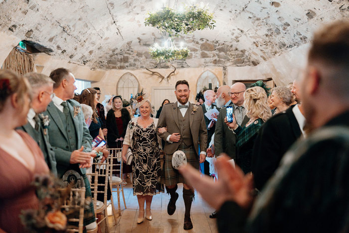 a groom wearing a kilt walking down an aisle on the arm of a woman wearing a black and white floral dress as rows of guests watch on