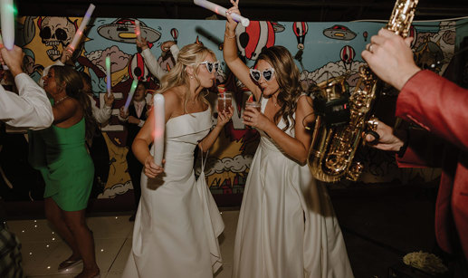 two brides wearing love heart sunglasses dancing with glow sticks on a light-up dance floor. They are both drinking cocktails through a straw. There is a saxophone in the foreground at the right hand side of the image