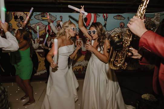 two brides wearing love heart sunglasses dancing with glow sticks on a light-up dance floor. They are both drinking cocktails through a straw. There is a saxophone in the foreground at the right hand side of the image