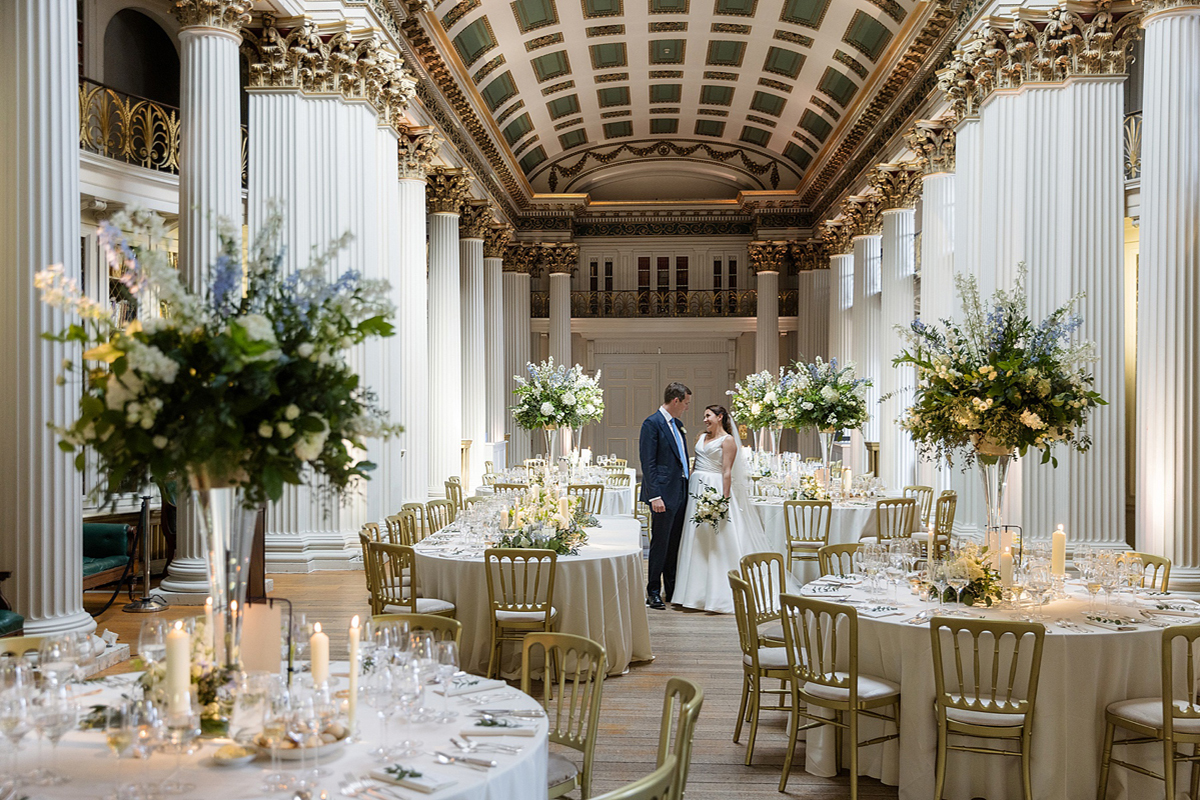 Bride and groom standing among elegant wedding tables in the Lower Library at the Signet Library, Edinburgh, with tall floral centrepieces and candlelight