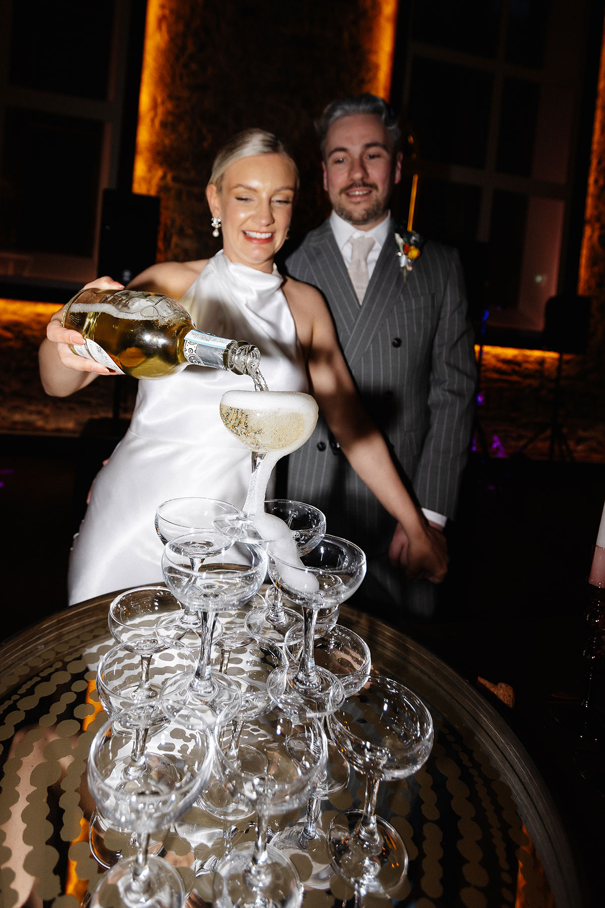 Bride pours champagne into a coupe tower as the groom watches during their Virgin Hotels Edinburgh wedding reception, captured by Steph Nicol Photography.