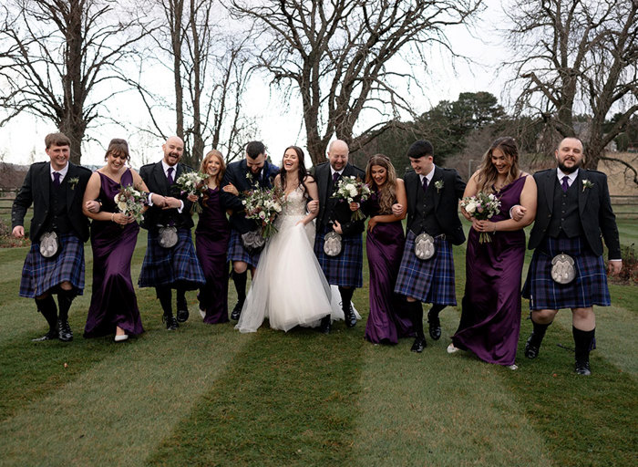 a line up of men in kilts, bridesmaids wearing purple dresses and a bride and a groom linking arms. They are walking across a lawn with bare trees in background.