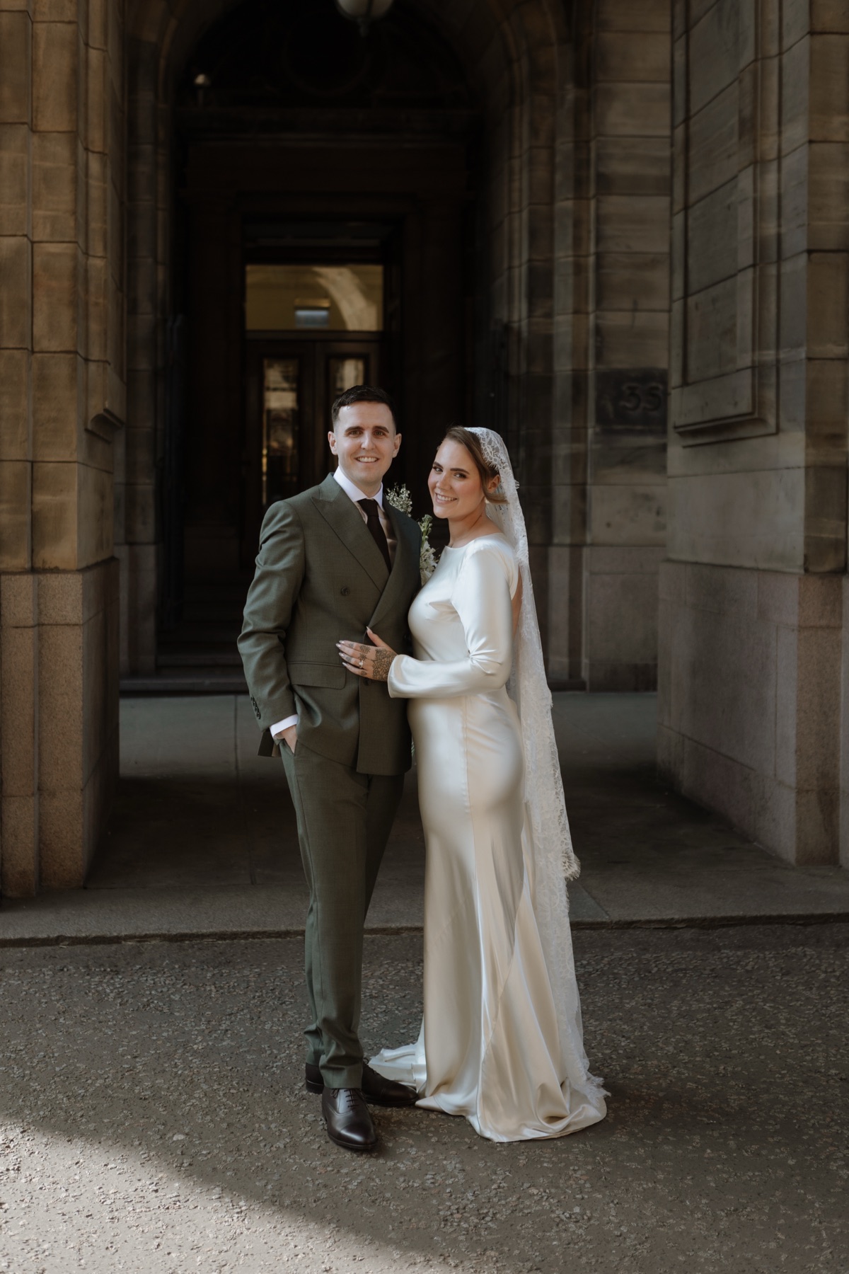 Bride and groom posing together beneath a stone archway during their Glasgow city wedding.