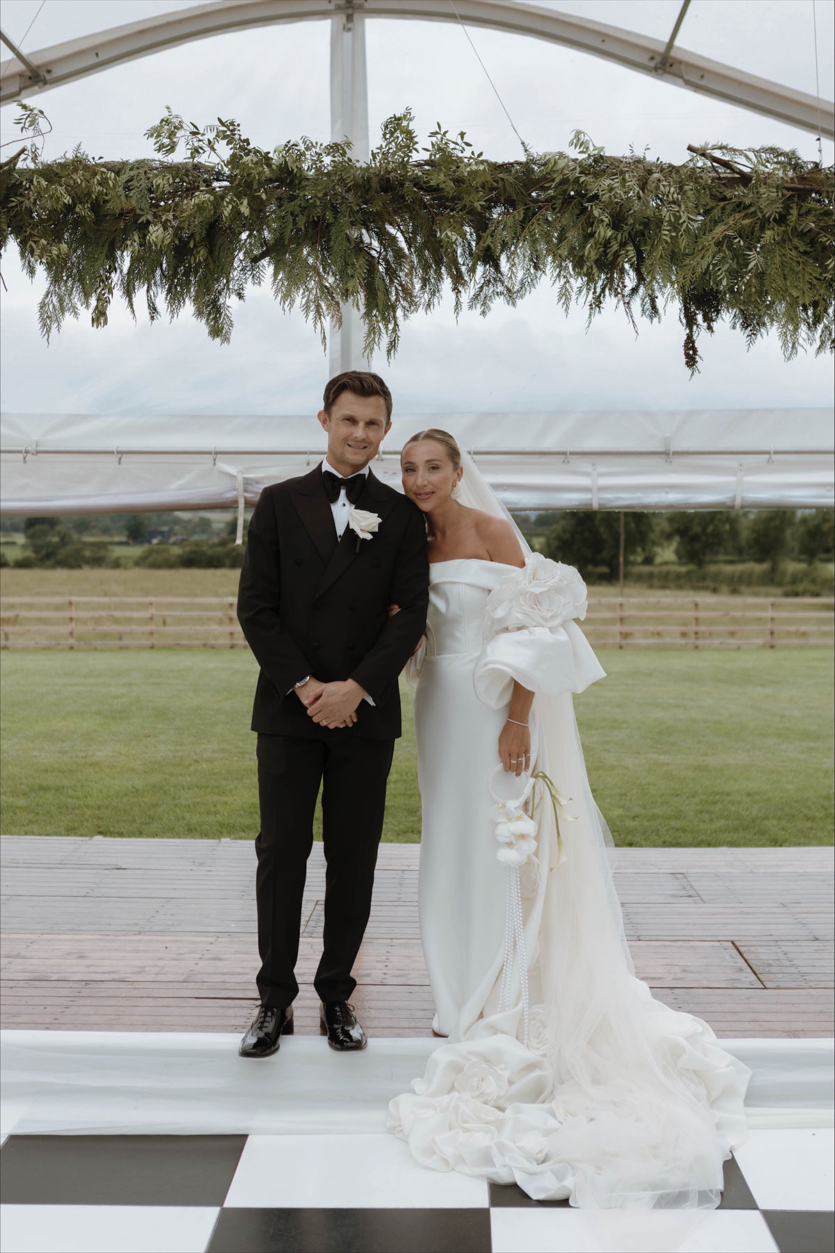 Bride and groom posing together in a marquee, bride wearing an off-shoulder gown and veil.