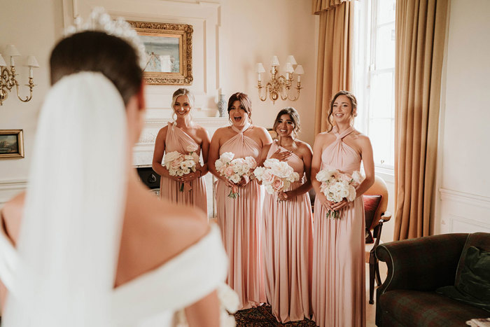 a bride faces away from the camera as the four bridesmaids wearing pink dresses look at her with surprised reactions 