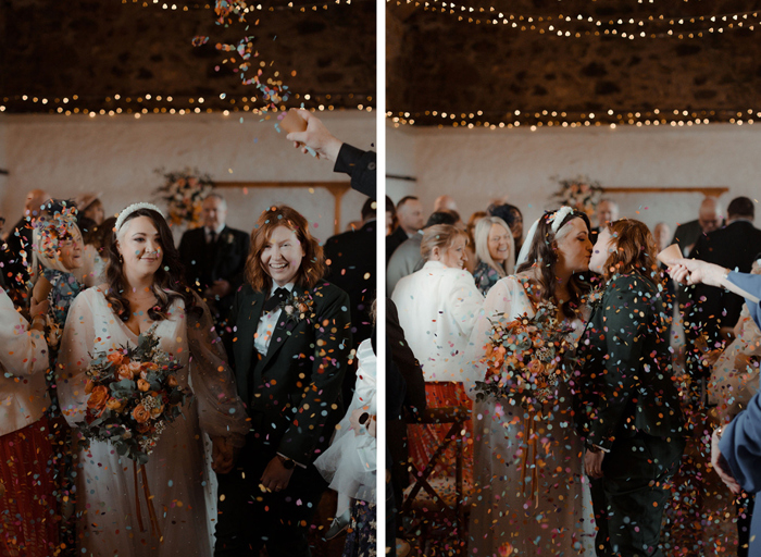 two brides, one wearing a wedding dress, one wearing a dark green suit, walking up the aisle while being showered in colourful rainbow confetti at the Cow Shed Crail