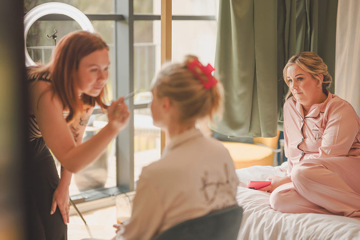 A woman applies makeup with a brush to a woman's face while another woman in pajamas looks on