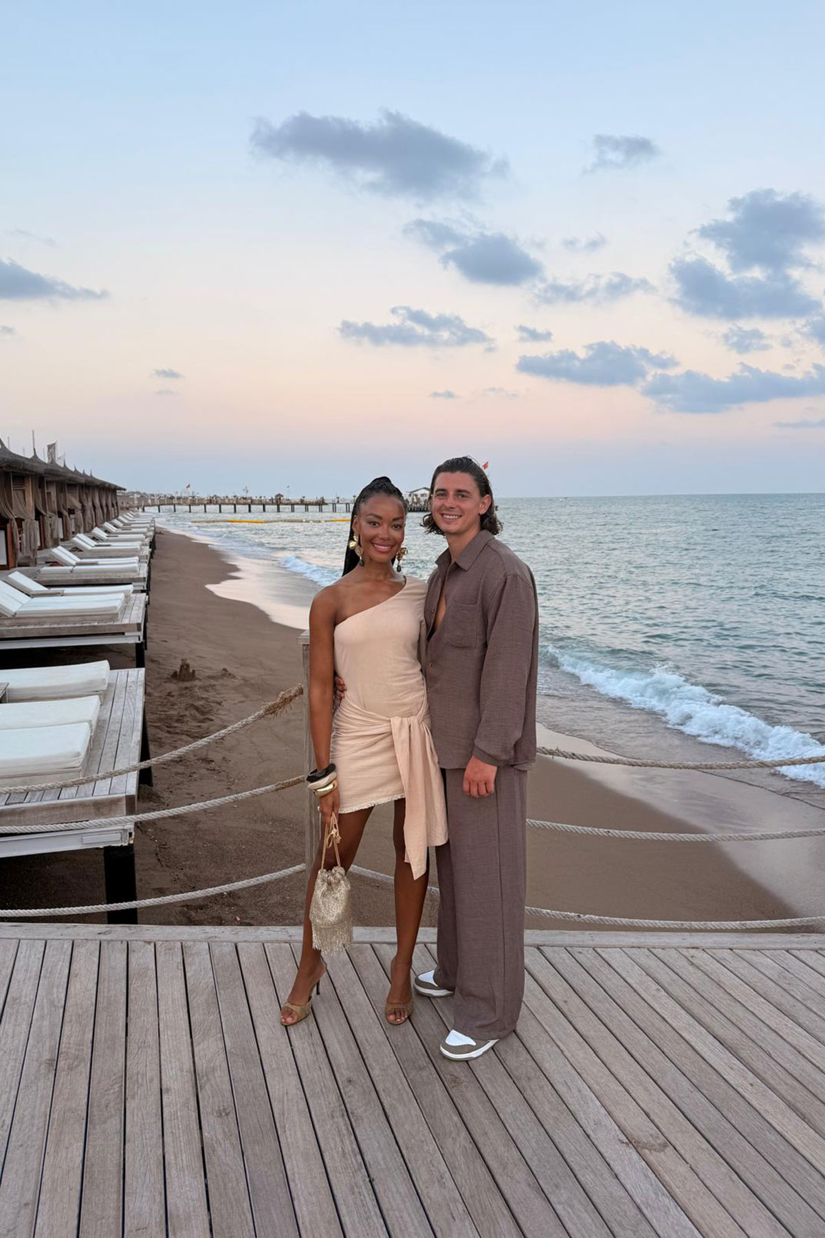 A couple stand arm in arm on a wooden boardwalk beside the beach, with soft evening light and waves behind them