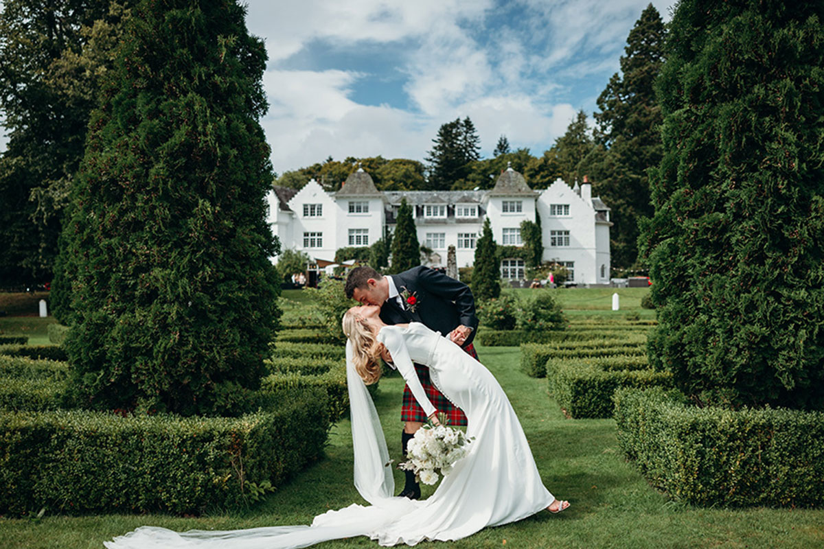 Bride and groom portrait in the formal gardens at Achnagairn Castle, with the couple sharing a kiss in front of the Highland estate's white façade and manicured hedges