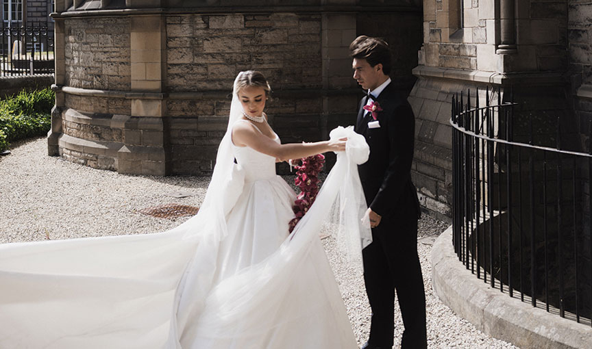 Bride twirling her long veil while standing with the groom outside the grand stone exterior of Mansfield Traquair.