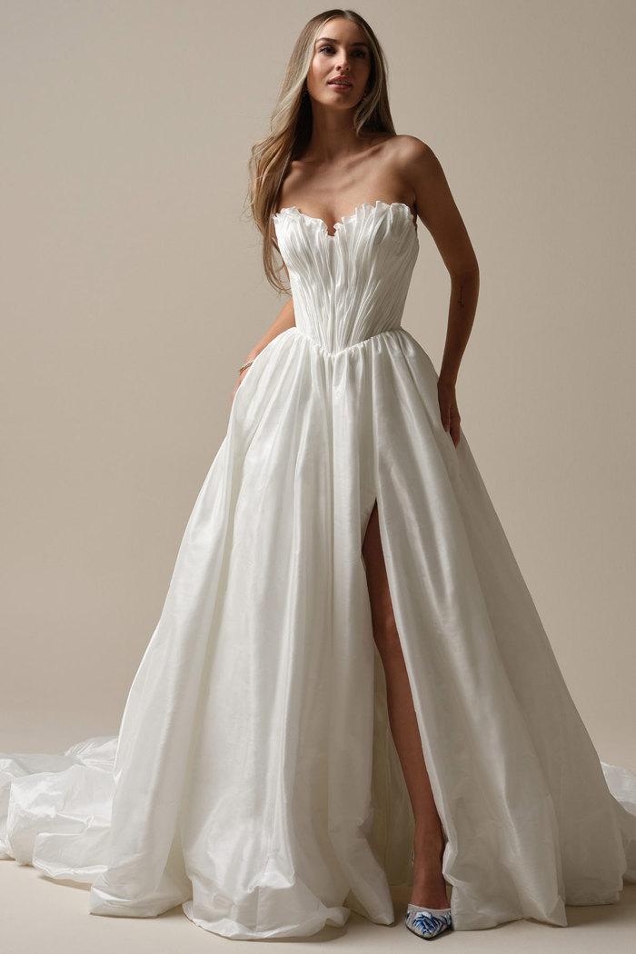 blonde model wears strapless wedding dress with ruffled bodice and large voluminous ballgown skirt with leg slit