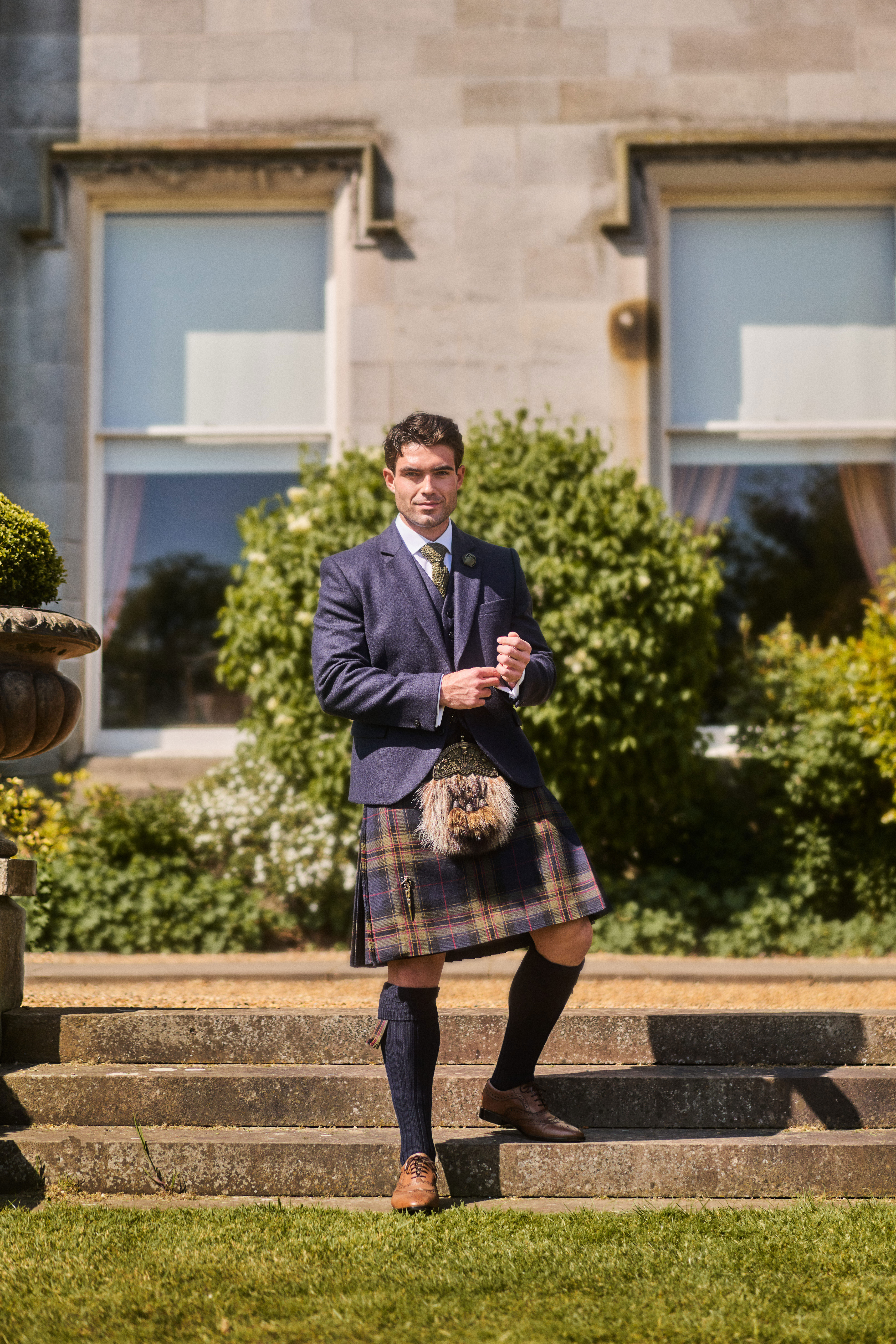 groom smiles while standing on steps at dundas castle as he wears mccalls highlandwear's new noble print tartan kilt
