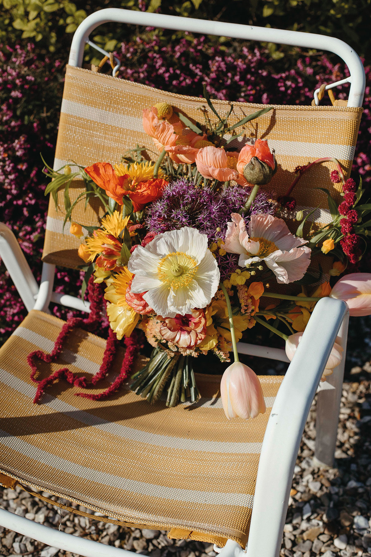 Bright, colourful wedding bouquet with poppies and seasonal blooms resting on a striped outdoor chair in the sunshine