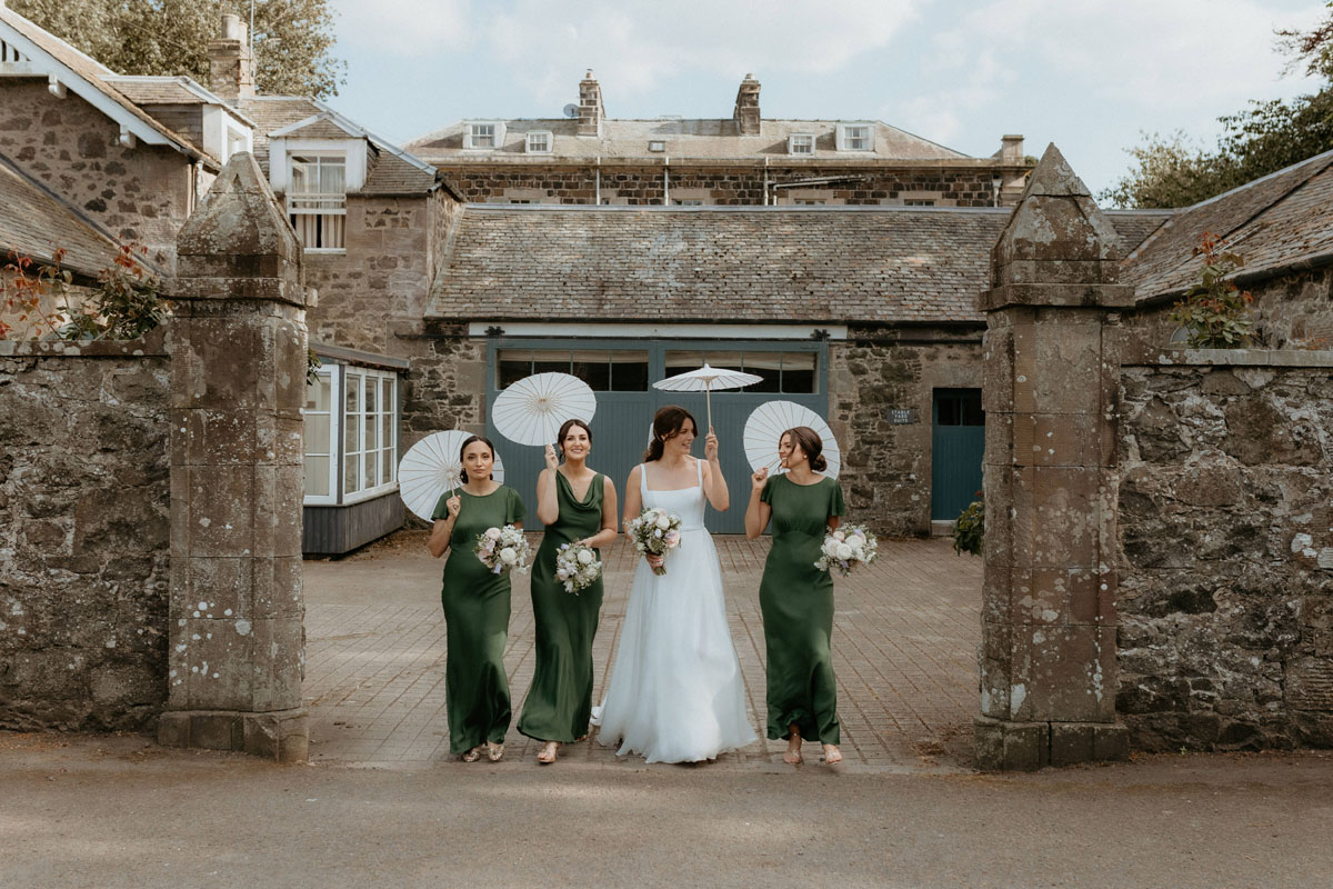 Bride and bridesmaids in green dresses carrying white parasols outside The Byre at Inchyra.