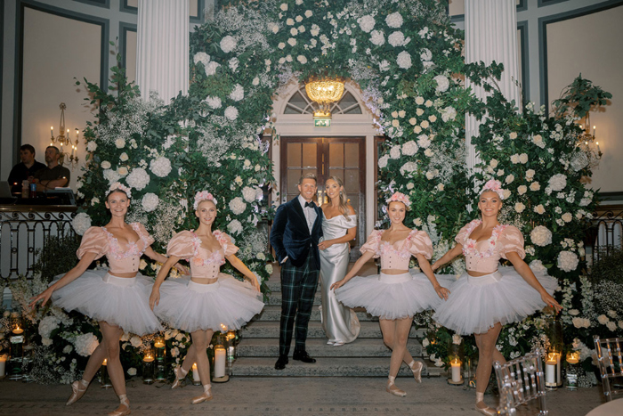 Bride and groom stand under floral arch with ballerina performers in front of them
