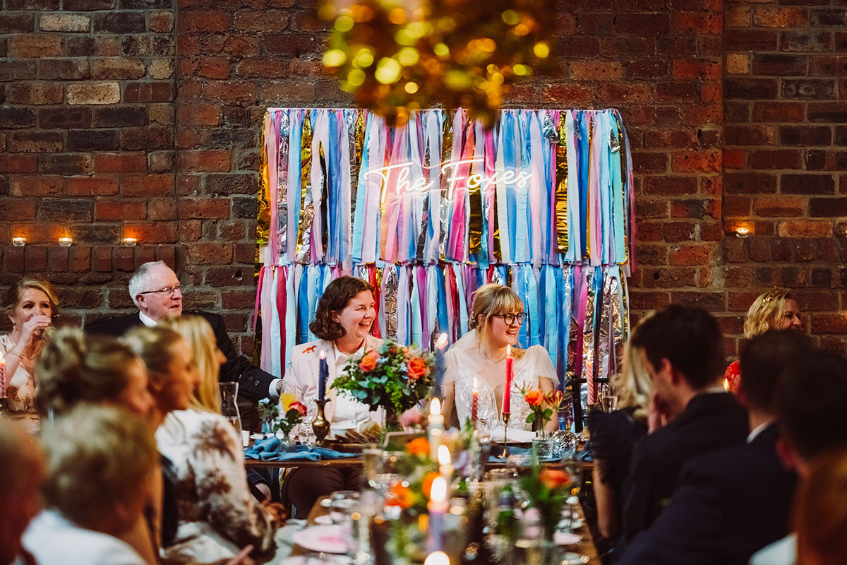 The newlyweds seated at their top table with a colourful streamer and neon sign backdrop during wedding speeches at the Engine Works.
