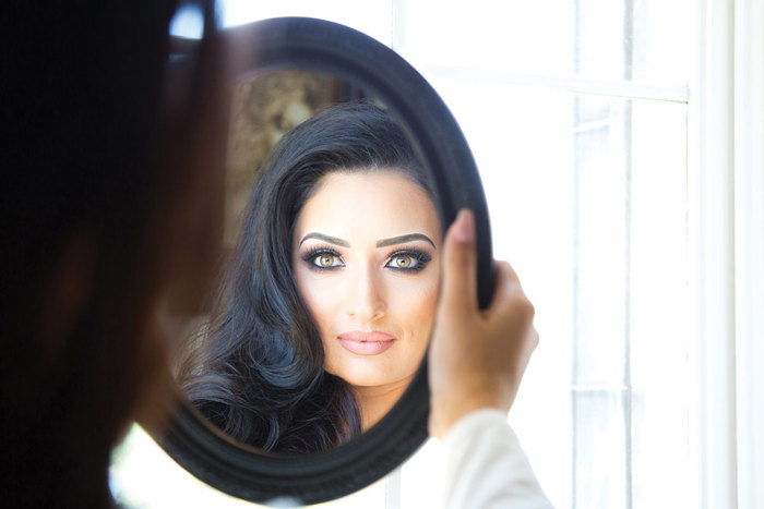 woman with long black hair and a full face of makeup looks into round handheld mirror