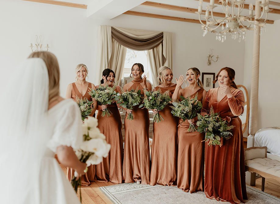 six bridesmaids wearing rust-coloured dresses look emotional and surprised as a bride in foreground holds bouquet. They are in an elegant room decorated in cream with light wooden details and draped curtains in the background