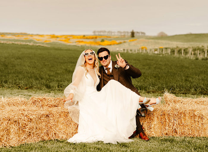 a bride and groom sitting on bales of hay in a field. They are wearing heart shaped sunglasses and the groom is making a peace sign with a hand while the bride laughs