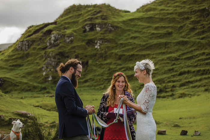 Couple tying ribbons together in handfasting ceremony near hills with celebrant watching on