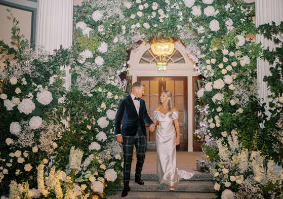 Bride and groom walk downstairs with white flower installation behind them