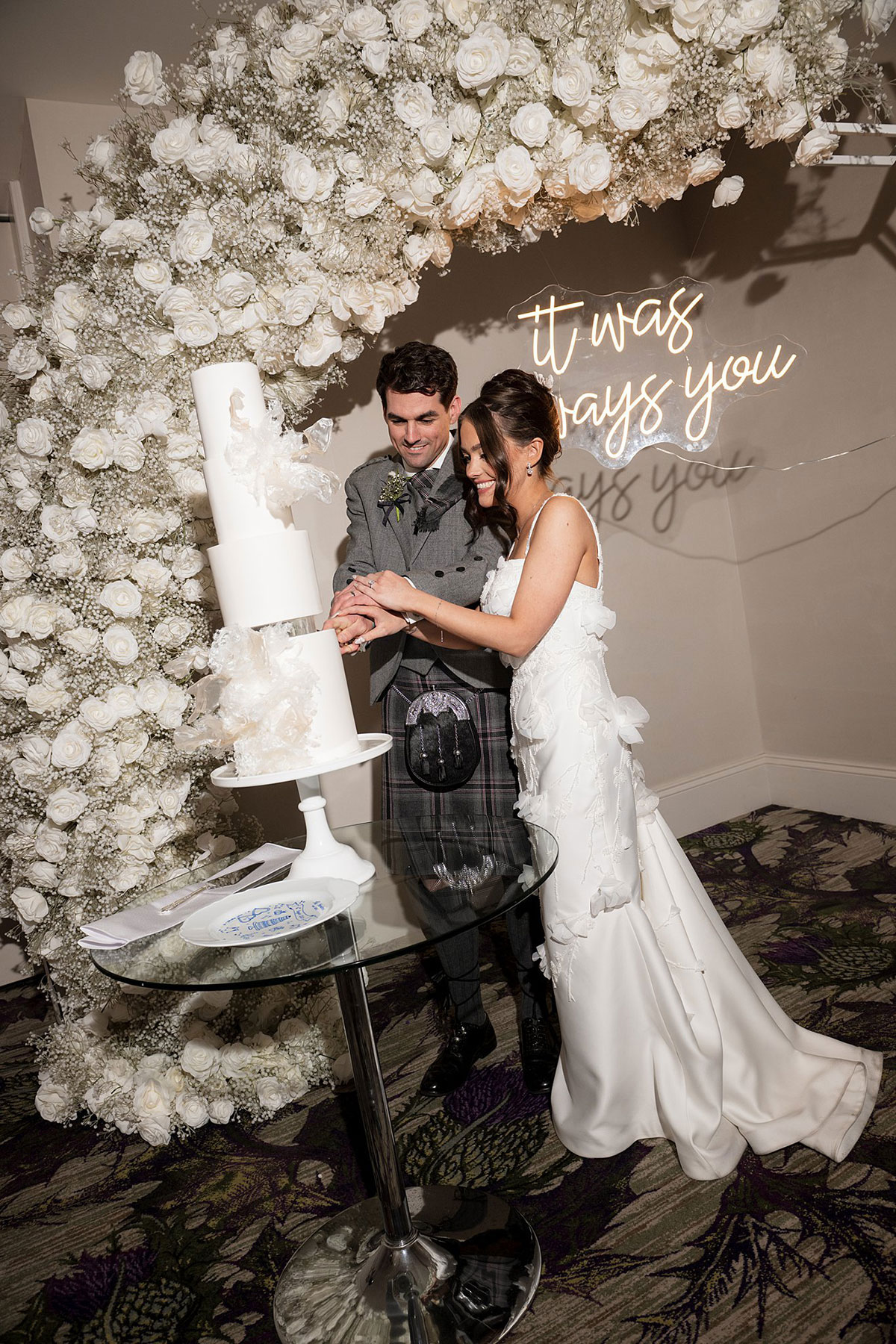 Bride and groom cutting their modern white wedding cake in front of a floral arch and neon sign at Old Course Hotel reception.