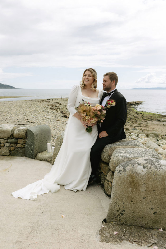 a bride perching on a groom's knee. They are sitting on a stone wall in front of a sandy and stony beach 