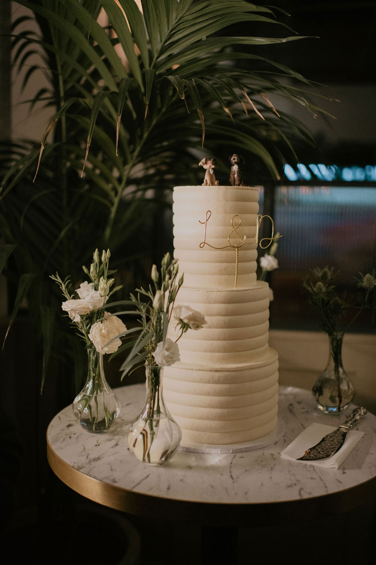 A three-tier wedding cake with the letters 'L & D' in gold stuck on and two fondant dogs on the top