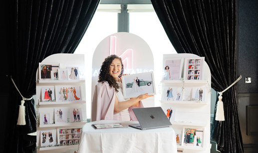 a brunette woman sits behind a table and macbook, smiling and surrounded by a display of printed out portraits