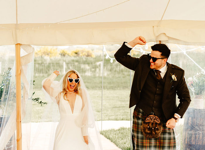 a bride and groom wearing heart shaped sunglasses cheer and raise one arm in the air with clear windows of a marquee behind them