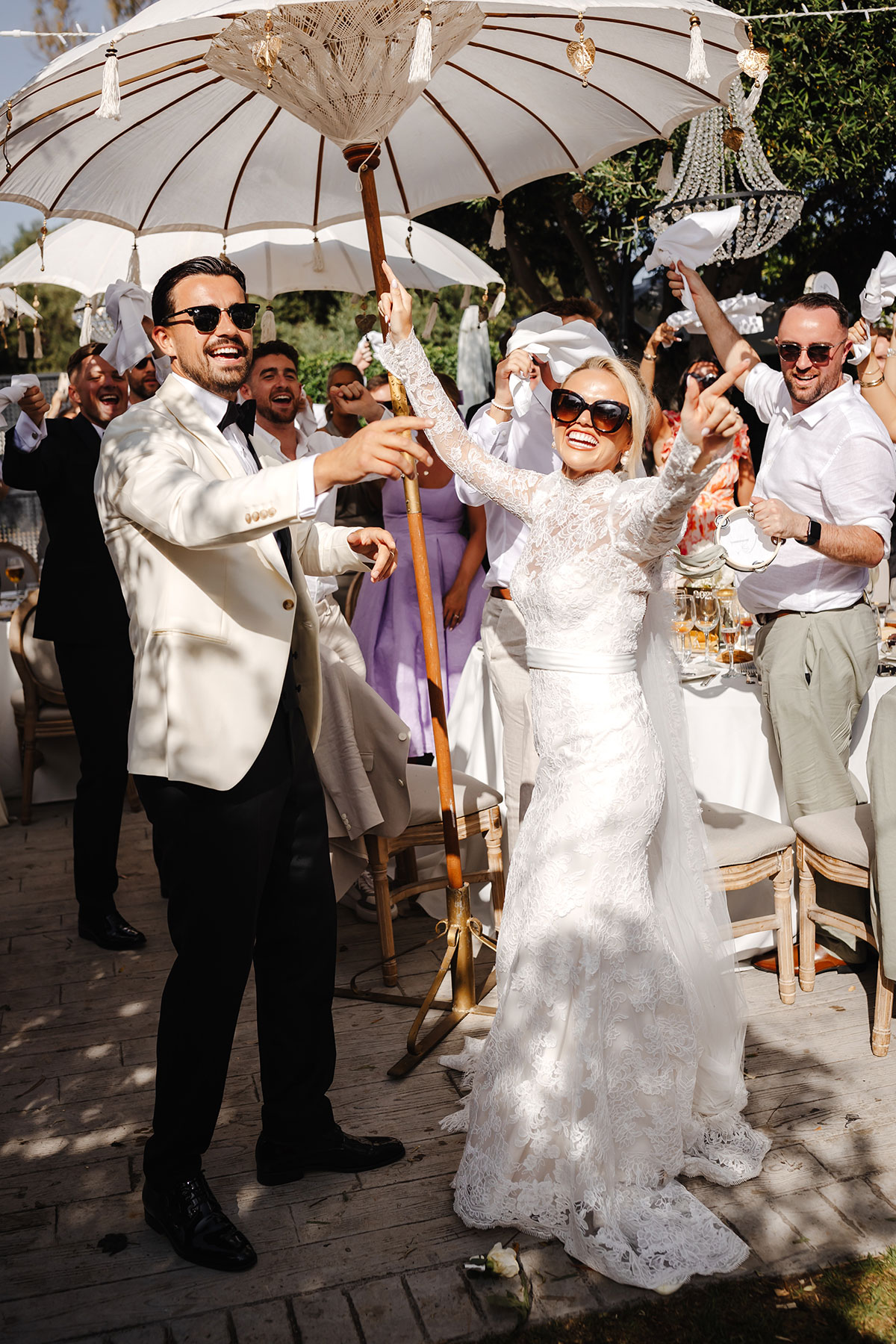 Bride and groom wave parasol surrounded by cheering guests at sunlit outdoor reception
