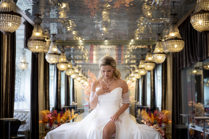 A woman wearing a strapless wedding dress with tulle sleeves and a slit in the skirt sitting on a table in a mirrored room with crystal chandeliers