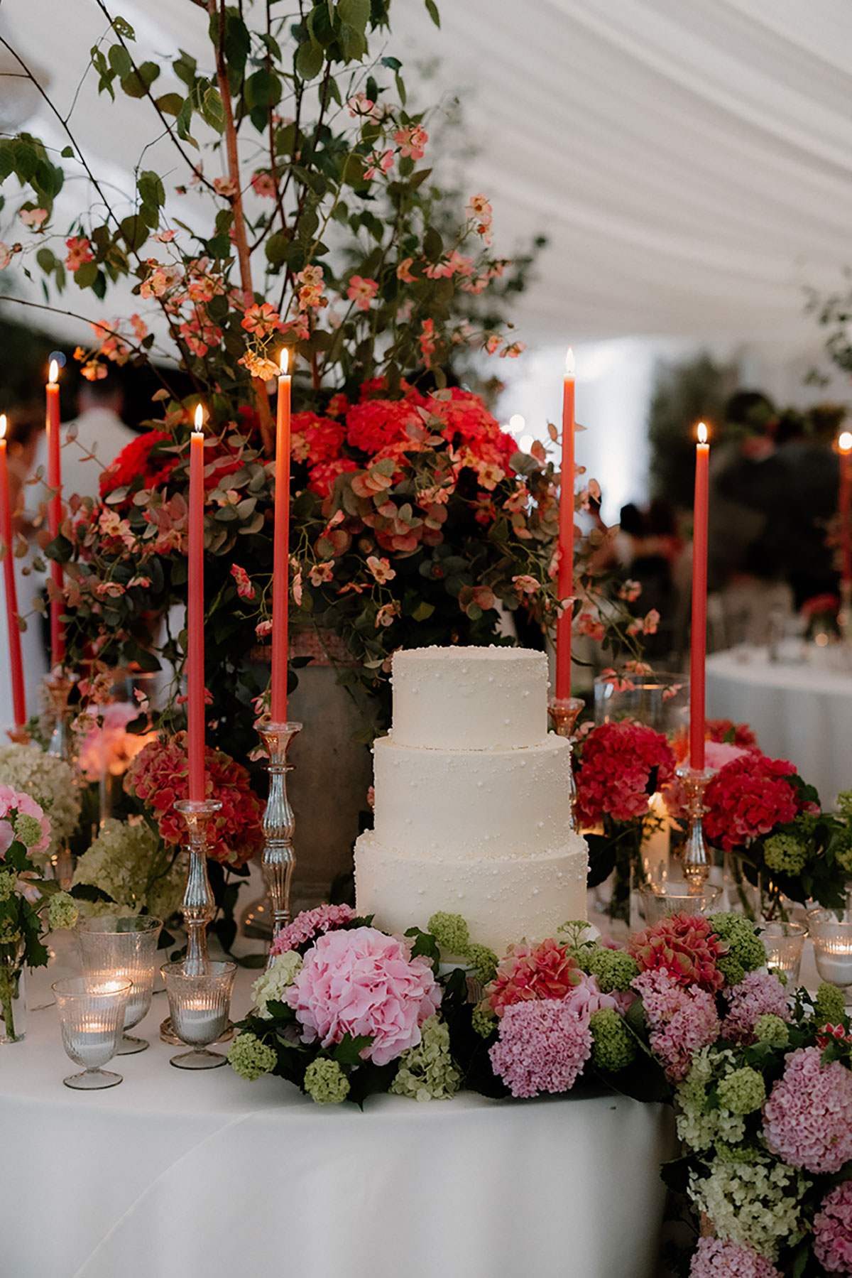 Three-tier white wedding cake surrounded by vibrant flowers and tapered candles in the reception marquee.
