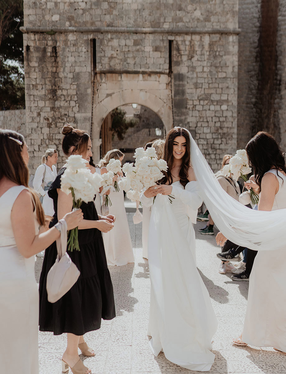 A bride in a white dress stands smiling, holding a bouquet of white flowers, surrounded by bridesmaids outside a stone building.