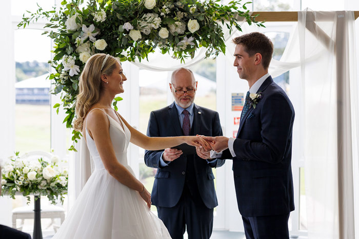 A joyful bride and groom exchanging rings during their Duke Photography captured wedding ceremony, with an officiant standing between them underneath a floral arch at the Old Course Hotel