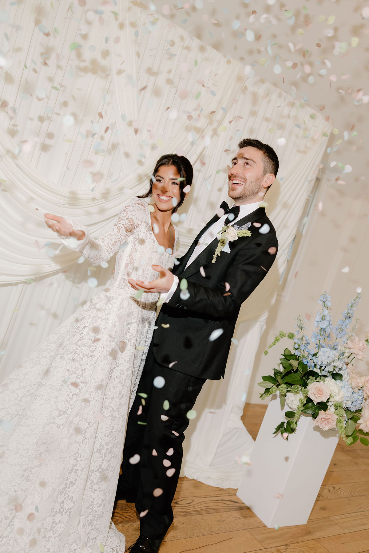 Bride and groom smiling beneath a shower of pastel confetti beside floral arrangements