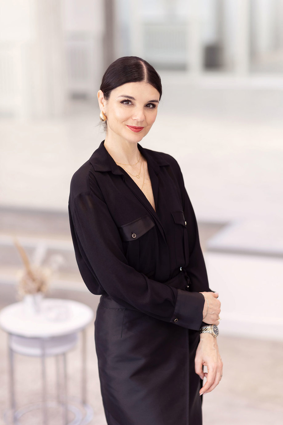 woman in sleek black outfit and and middle part stands smiling softly with a red lip