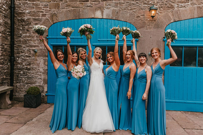 A bride in a white dress and seven bridesmaids in long blue dresses stand with their bouquets held above their heads