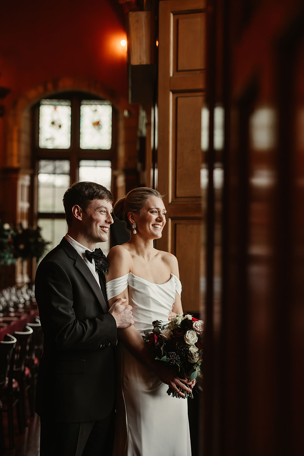 Bride and groom standing together in the candlelit Minstrels’ Hall, looking out towards the windows.