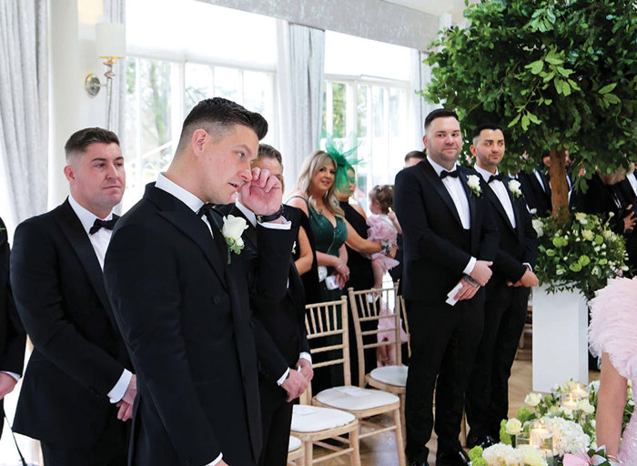 groom wearing a black suit and bow tie dabbing a tear away as other guests stand looking on