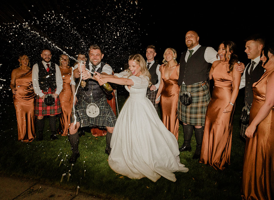 a bride and groom doing a champagne spray at each other in the dark with men in kilts and women in rust coloured bridesmaid dresses cheer and look surprised in the background