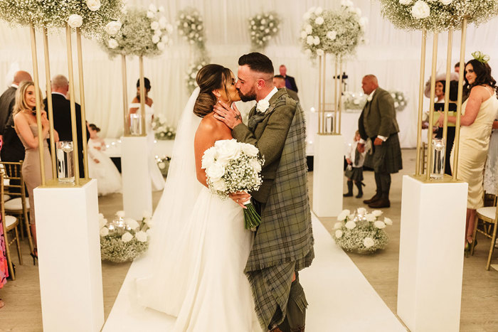 bride and kilt-wearing groom kissing in a venue decorated with white florals