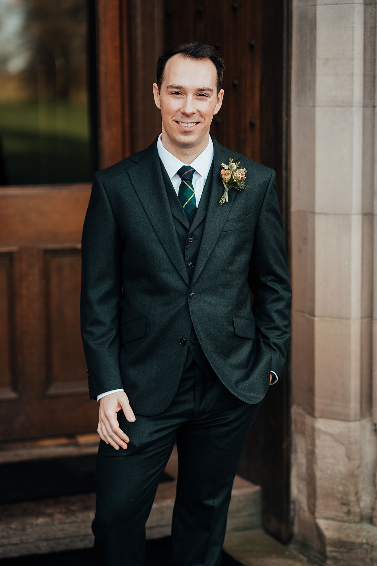 Groom in a dark three-piece suit with tartan tie and buttonhole standing outside Schloss Roxburghe