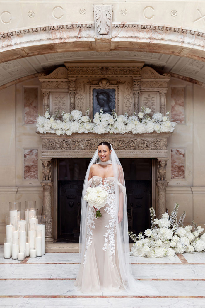 A bride in a Berta Privee wedding dress from Opus Couture standing in front of an ornate stone fireplace that's decorated with white flowers and candles.