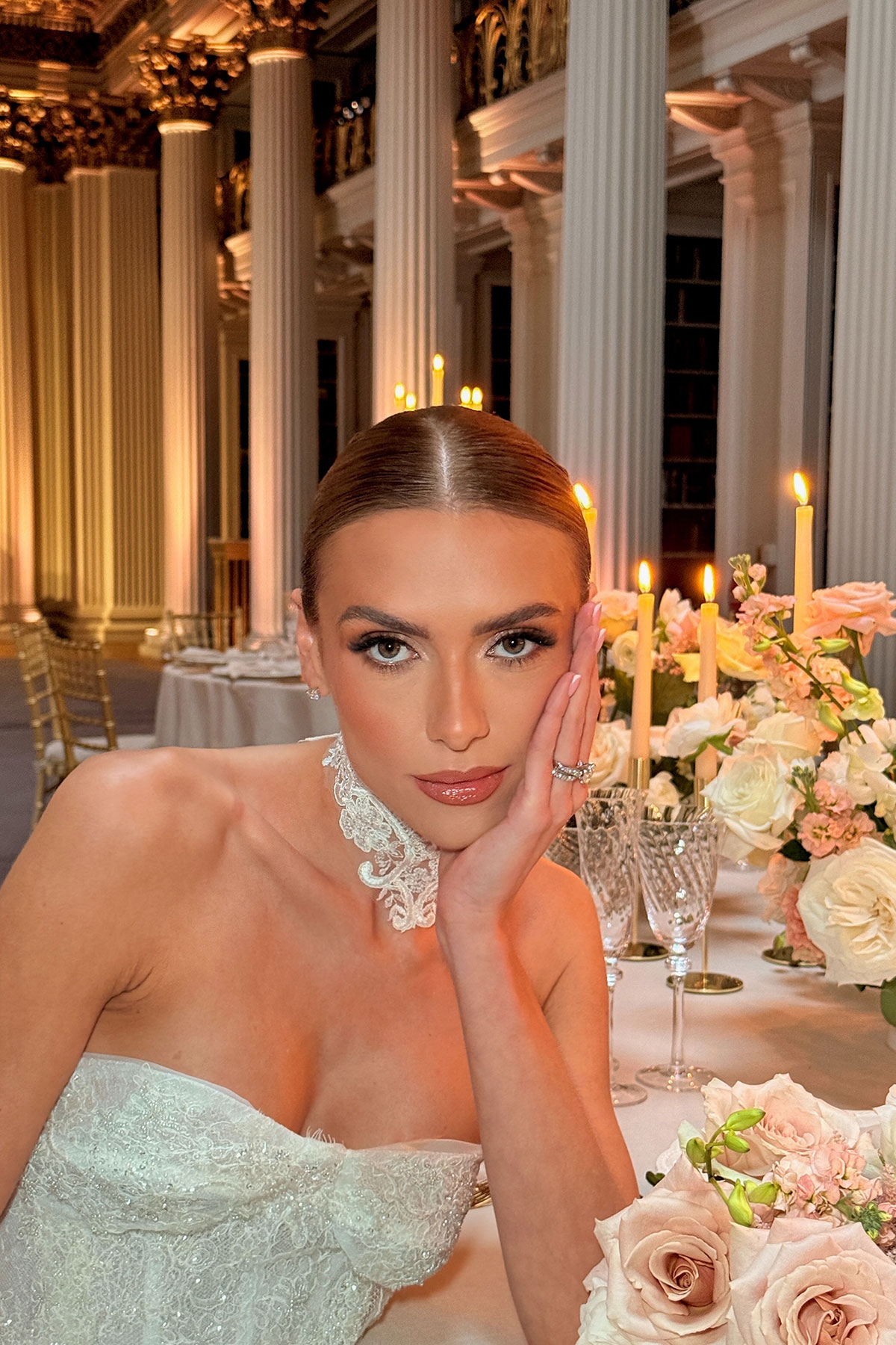 Bride with sleek centre-parted hairstyle and glowing makeup seated at an elegant candlelit reception table with pastel florals