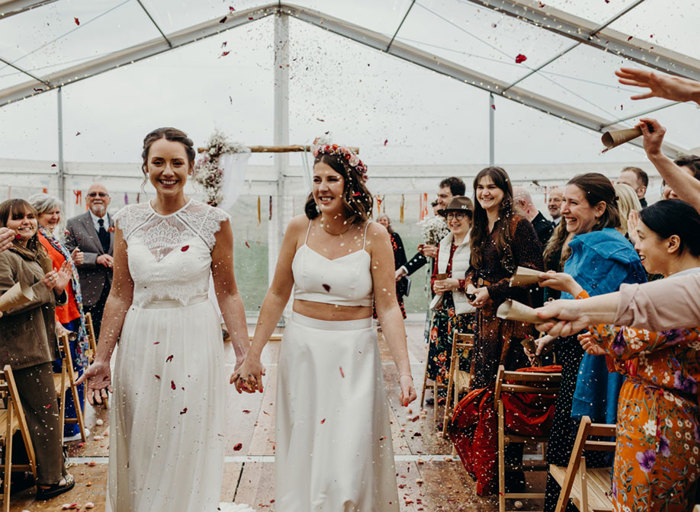two bride being showered in colourful confetti by wedding guests either side, while walking on a wooden floor in a marquee