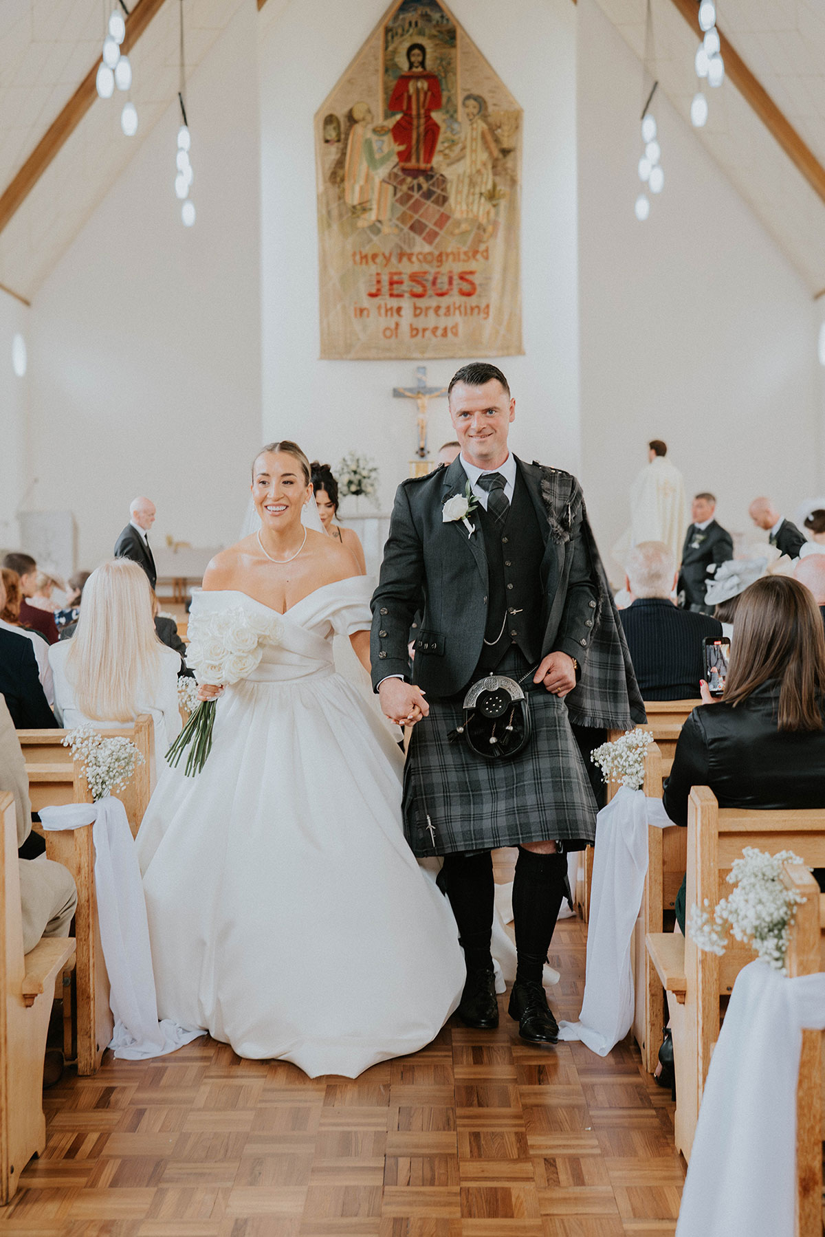 Bride and groom walking down church aisle after ceremony, bride holding white rose bouquet and groom in Scottish kilt