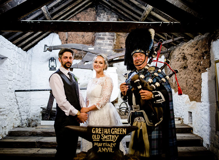 a bride and groom standing with a bagpiper in an old stone barn with whitewashed walls and exposed wooden beams
