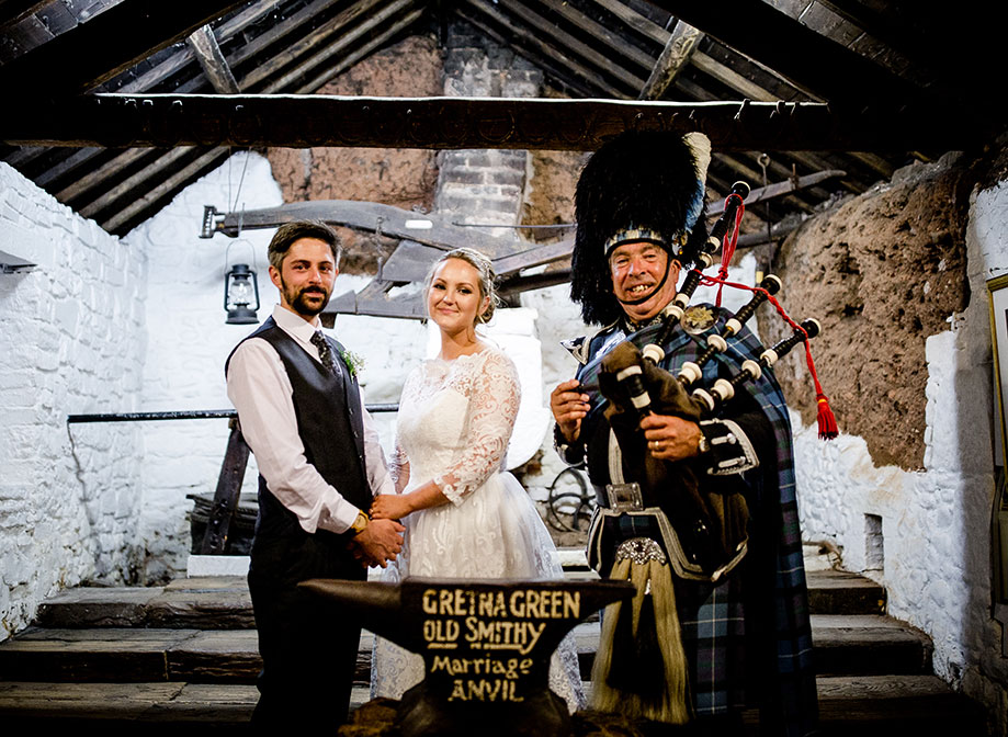 a bride and groom standing with a bagpiper in an old stone barn with whitewashed walls and exposed wooden beams